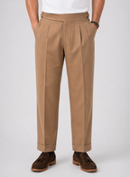 Men's tan relaxed twin-pleat high-waist pants with side adjusters and cuffed hems, paired with brown loafers.