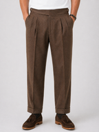 Men's brown relaxed twin-pleat high-waist pants with side adjusters, cuffed hems, and brown suede loafers.